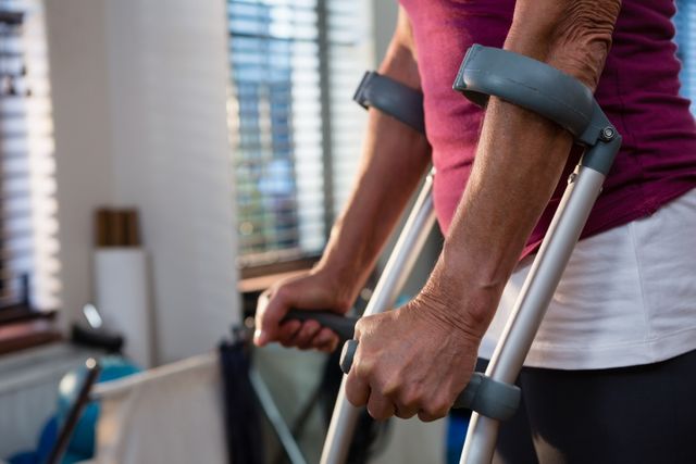 This image shows a close-up of a woman using crutches in a clinic, emphasizing her hands and the crutches. Ideal for use in healthcare, rehabilitation, and physical therapy contexts, it highlights themes of recovery, support, and mobility aid. Suitable for articles, blogs, and promotional materials related to medical care, patient recovery, and senior healthcare.