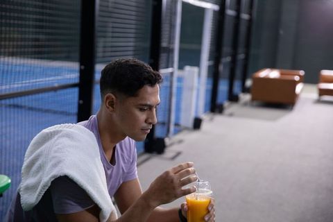 Indian athlete resting on bench holding orange juice after match beside blue indoor court