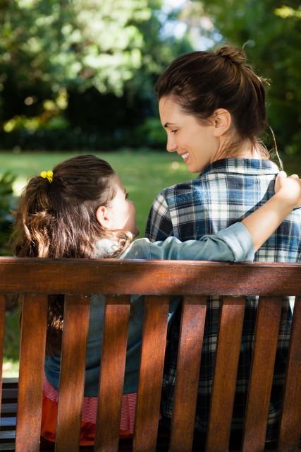 Smiling woman looking at daughter with arm around on wooden bench at backyard