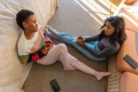 African american and indian women sitting on floor chatting over coffee in cozy sunny living room