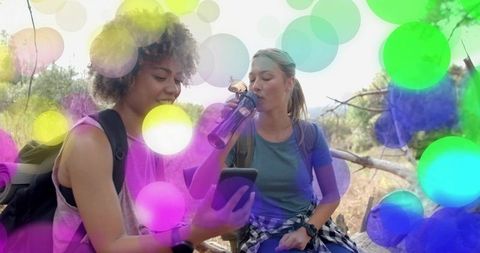 Two female hikers resting on log checking phone and drinking water with colorful bokeh