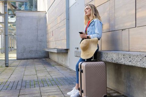 Young woman leaning on urban bench waiting with suitcase and sunhat checking smartphone