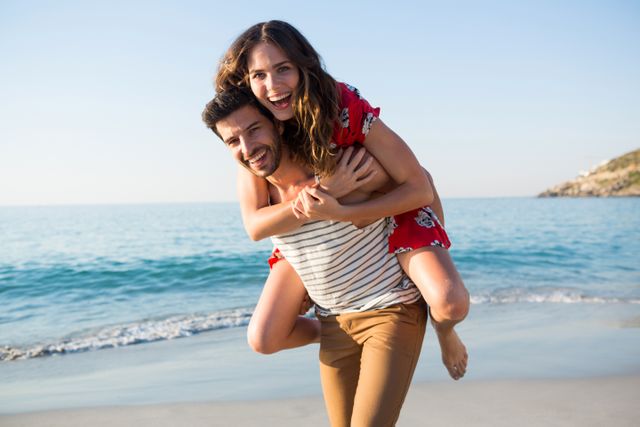 Portrait of young man piggybacking his cheerful girlfriend at beach on sunny day