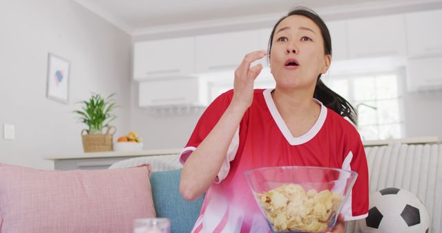 Image of exited asian woman eating chips and watching match in tv. Family, motherhood, relations and spending quality time together concept digitally generated image.