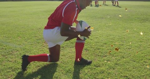 Kneeling rugby player holding ball on sunlit grass pitch with gold star highlights