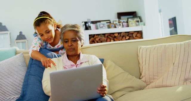 Grandmother sitting on sofa with granddaughter using laptop, indicating family bonding through technology and learning. Useful for topics on intergenerational connections, technology use by seniors, family life, and domestic settings.