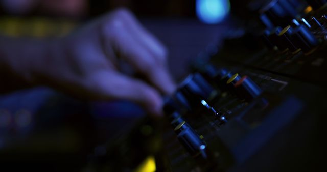 Close-up view of a person's hand adjusting controls on an audio mixer in a low light environment. Ideal for use in publications about music production, audio technology, sound engineering, professional studio settings, and concert management. Can be utilized for blogs, articles, advertisements, and promotional materials related to studio equipment or sound technology.
