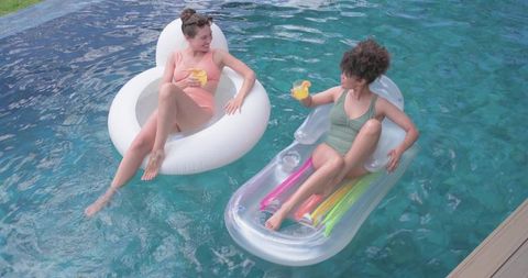 Women relaxing on inflatable pool floats holding drinks enjoying sunny summer poolside leisure