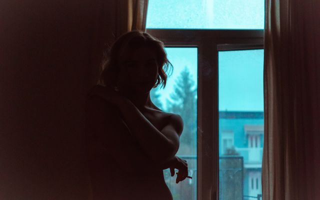 Silhouette of a woman standing by a window with a blue sky visible outside. The scene creates a mood of contemplation and serenity. This composition can be used for themes of introspection, solitude, or minimalistic design. Ideal for use in articles or projects focusing on mental health, relaxation, and quiet moments.
