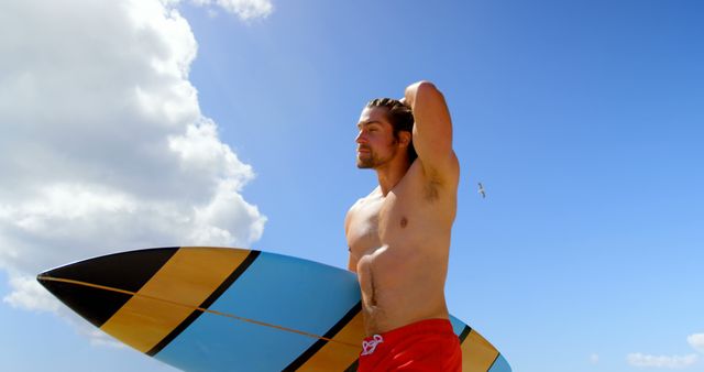 A young man stands on a sunny beach holding a surfboard, looking towards the ocean. He is shirtless and wearing red swim trunks, showcasing a fit and athletic physique. The blue sky is dotted with a few clouds, and a seagull is visible in the background. Suitable for portraying outdoor activities, beach lifestyle, fitness, leisure, and summer vacation themes.