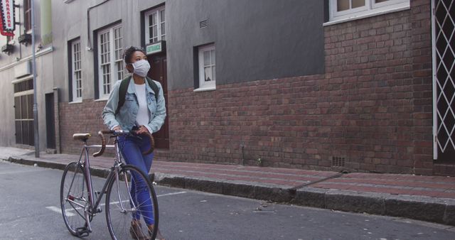 Happy asian woman wearing face mask wheeling bicycle in city street, looking at buildings. City living, travel and healthy modern urban lifestyle.
