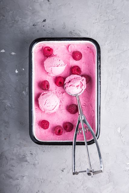 A tray of pink raspberry ice cream with a metal scoop taken from above. Additional raspberries decorate the ice cream. Ideal for food blogs, recipe sites, or ads for dessert or dairy products. Perfect for summer-themed content or featuring homemade and gourmet dessert ideas.