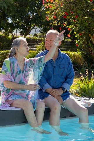 Diverse senior couple sitting at pool edge dipping feet and pointing at red blooms