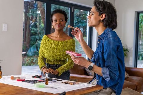 Makeup artist applying blush to woman in green textured top in bright modern living room
