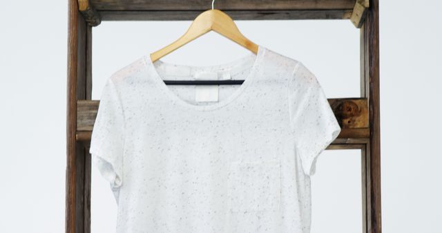 A plain white speckled t-shirt hangs on a wooden hanger against a neutral background, with copy space. Its simplicity suggests a minimalist fashion style or a blank canvas for customization.