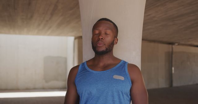 Young muscular African American man meditating under a large concrete structure while wearing a blue sportswear tank top. Useful for themes focused on fitness, mental health, urban lifestyle, mindfulness, and personal wellness. Ideal for posters, brochures, websites, and articles promoting meditation, relaxation techniques, and balanced lifestyles.