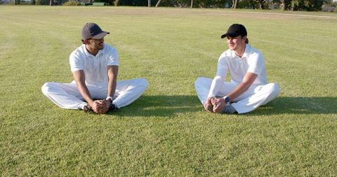 Stretching and chatting cricket teammates on sunlit grass field wearing white kit and black caps
