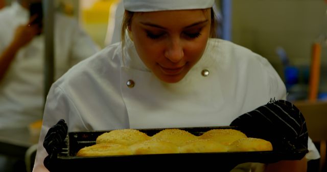 Woman baker in a commercial kitchen holding a tray of freshly baked buns and enjoying their aroma. This image can be used for bakery advertisements, culinary blog posts, recipes, or promoting baking classes and routines.