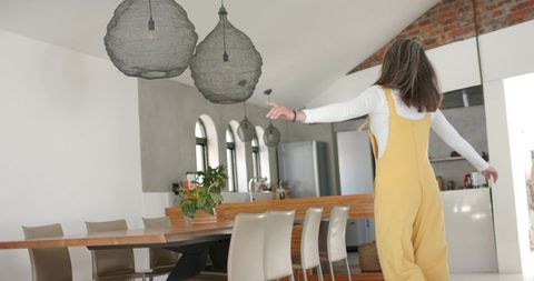 Mature woman twirling in sunlit open-plan kitchen wearing yellow overalls and white top