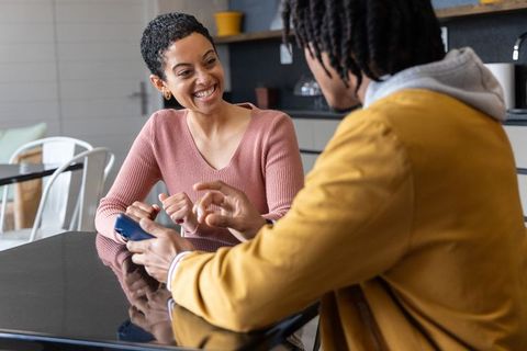 African american couple smiling and sharing smartphone at glossy kitchen table