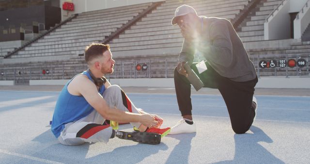 An athlete with a prosthetic leg sits on a track field while tying his shoes, with a coach kneeling beside him giving advice. This image can be used for themes related to sports, motivation, training, teamwork, coaching, determination, inclusivity, and disability sports. Suitable for use in articles, blogs, sports event promotions, motivational and inspirational campaigns, and informational content on adaptive sports or coaching techniques.