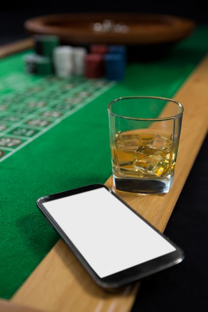 This image shows a mobile phone and a glass of whisky placed on a roulette table in a casino. The green felt and betting chips in the background suggest an active gambling environment. This image can be used for themes related to gambling, nightlife, technology integration in casinos, or luxury leisure activities.