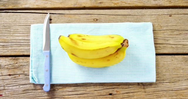 A bunch of ripe bananas lies next to a knife on a striped cloth over a wooden surface, with copy space. Bananas are a nutritious fruit, and this setup suggests preparation for a healthy snack or meal ingredient.