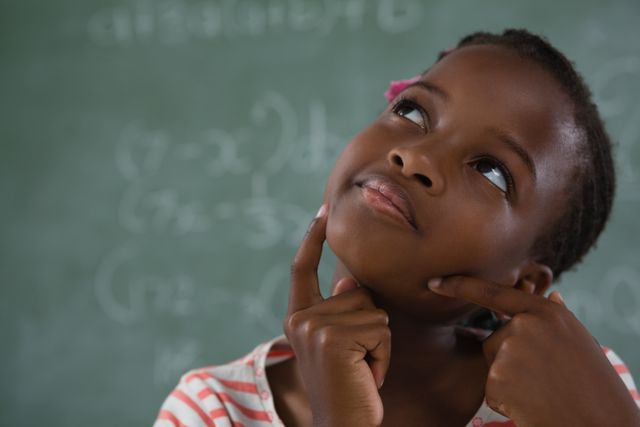 Young schoolgirl in striped shirt thinking deeply while sitting in front of chalkboard with mathematical equations. Ideal for educational content, school-related articles, and advertisements promoting learning and curiosity in children.