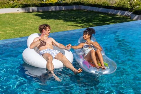 Mixed-race couple relaxing on inflatable floats in backyard pool holding hands with drinks