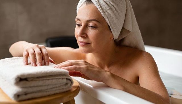 Middle-aged woman relaxing in bathtub reaching for folded towels with towel on head