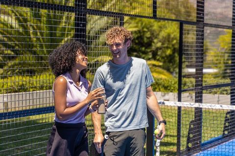 Couple enjoying padel match on sunny outdoor court holding paddle and water bottle