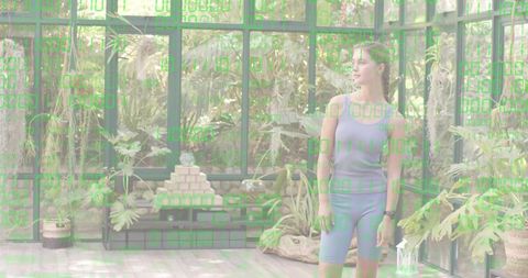 Woman standing in glass conservatory in athletic tank and shorts, gazing at lush indoor garden