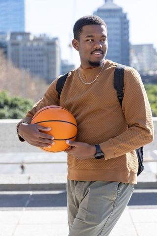 African american man holding basketball in urban plaza wearing backpack, casual sweatshirt