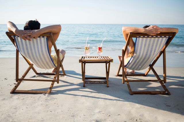 Couple enjoying a sunny day at the beach, sitting on lounge chairs with refreshing drinks on a small table between them. Ideal for use in travel brochures, vacation advertisements, and lifestyle blogs promoting relaxation and beach destinations.