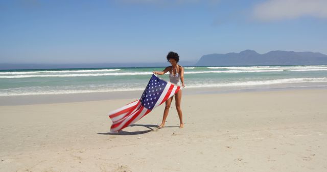 A young African American woman is walking along a sandy beach holding an American flag, with copy space. Her expression of joy and pride suggests a celebration of national spirit or a patriotic holiday.