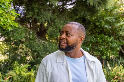 Bearded man standing in sunlit park looking left among lush green foliage