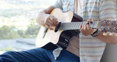 Middle-aged man playing acoustic guitar on sunlit balcony enjoying leisure