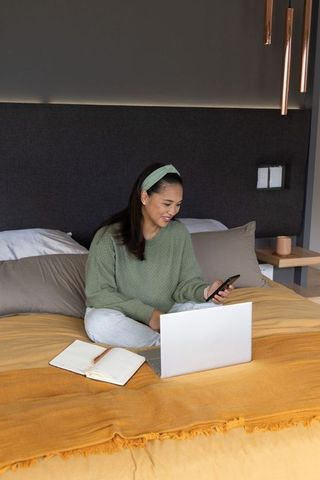 Asian woman working remotely on laptop and phone in cozy modern bedroom, studying and planning