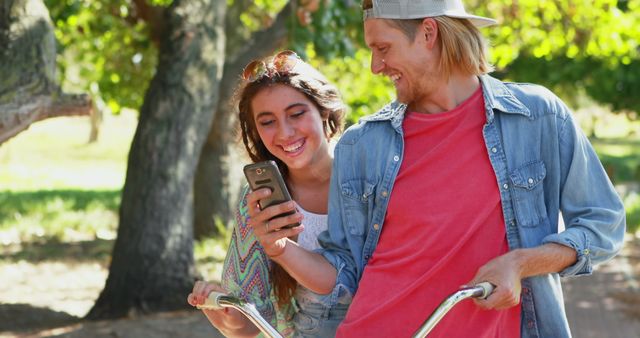 Young couple is enjoying a bike ride in a park while sharing and looking at something on a smartphone, both smiling and engaging with each other. Photo is ideal for promoting leisure activities, outdoor fun, friendship, and technology-related themes. Great for illustrating social media moments, lifestyle blogs, and advertisements catering to young adults.