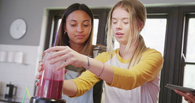 Two teenage girls are preparing smoothies together in a modern kitchen. One is operating a blender filled with red liquid, while the other watches closely. The environment is casual and friendly, promoting themes of healthy living, cooperation, and friendship. This image is ideal for use in health and wellness content, cooking blogs, or advertisements targeting young people and healthy eating habits.