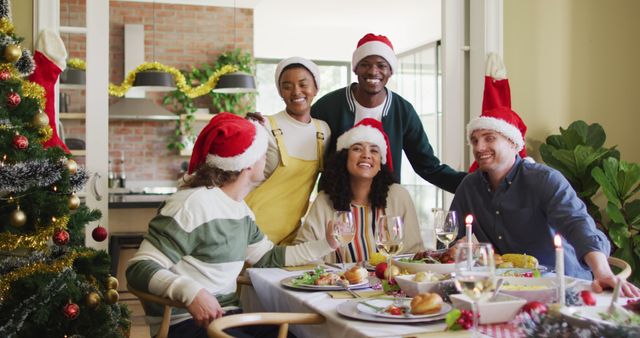 Group of friends celebrating Christmas dinner at home, wearing Santa hats and smiling around table. Perfect for holiday marketing campaigns, festive promotions, social media content, and advertisements promoting diversity and togetherness during holiday season.