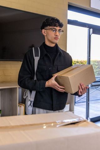 Asian man holding sealed cardboard box in modern lobby, wearing glasses and backpack