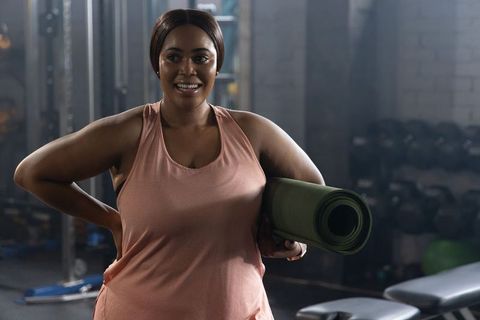 Confident african american woman holding rolled green yoga mat smiling in gym wearing dusty pink tan