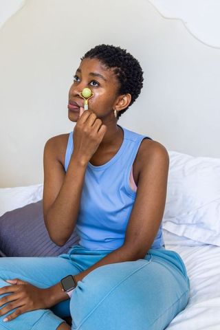 Young woman using jade facial roller under-eye sitting on bed pastel wellness routine