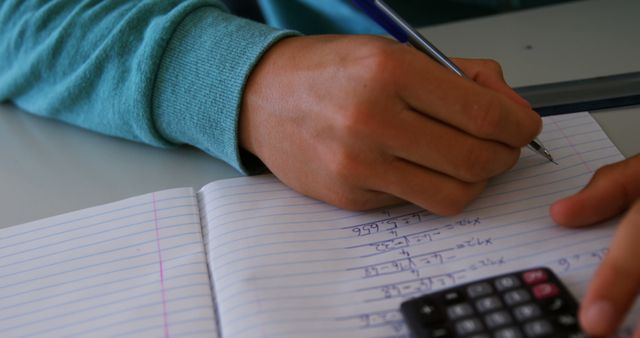 Person writing math problems in a notebook with calculator nearby. Useful for education, studying, mathematics websites and school-related content.
