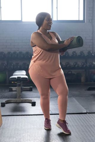 African american plus size woman holding green yoga mat in gym wearing peach activewear