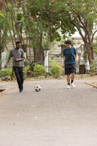 Multiracial friends walking and playing soccer on suburban tree-lined driveway
