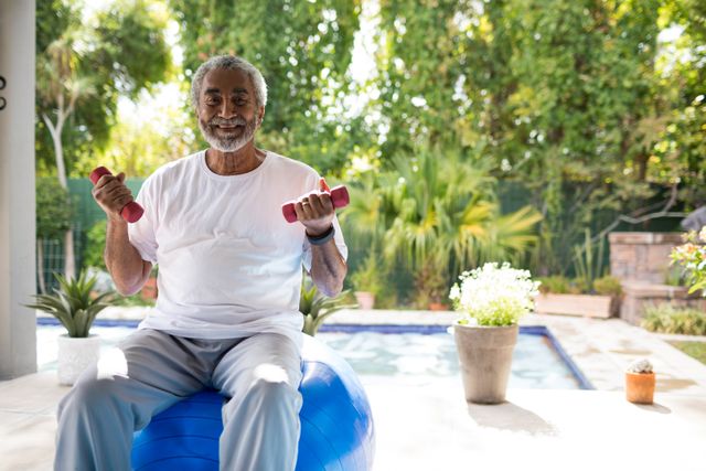 Portrait of man holding dumbbells while sitting on fitness ball in yard