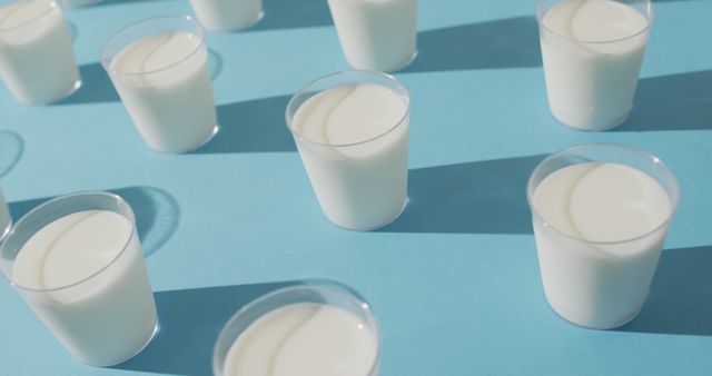 This image shows multiple small glasses of milk arranged on a blue surface, casting distinct shadows. It is ideal for concepts related to nutrition, dairy products, health beverages, and dietary calcium. It can be used in advertisements for dairy products, health blogs, nutrition articles, and social media campaigns focusing on promoting healthy eating habits.