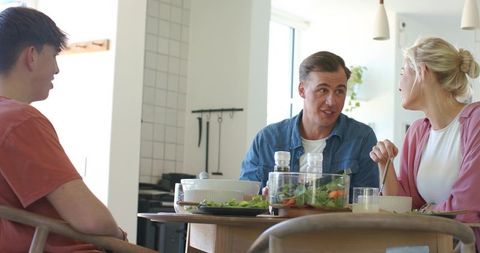 Three friends sitting at dining table sharing salad and chatting in bright home kitchen